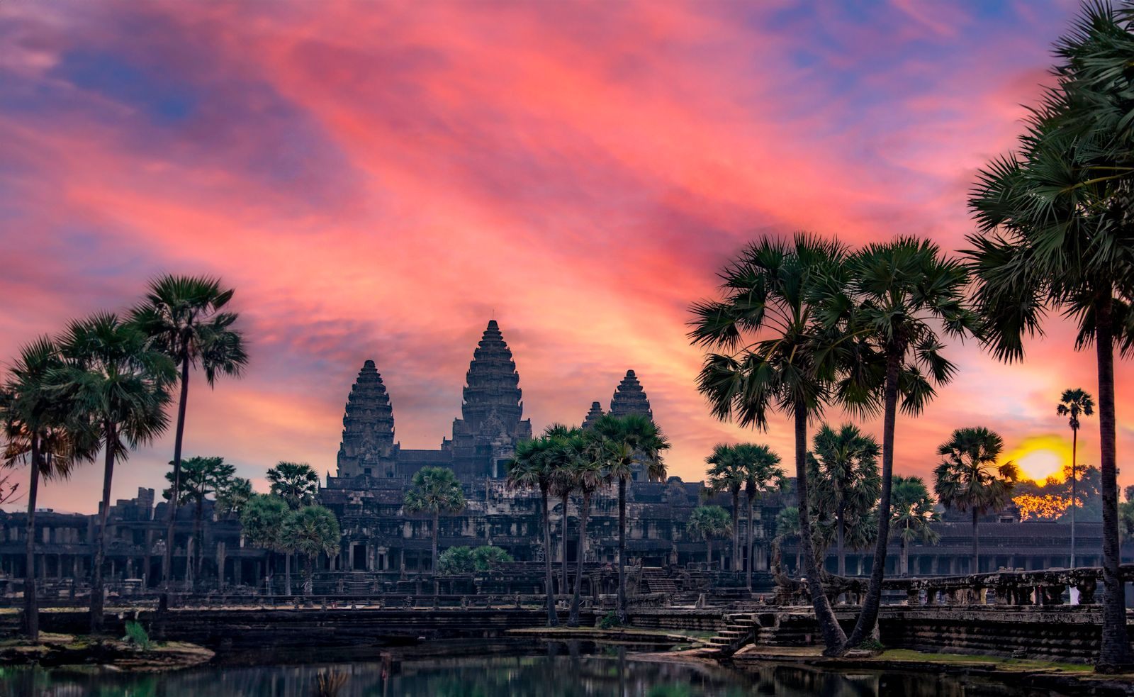Towers of Angkor Wat silhouetted by palms under a red sunset sky, a favorite Mekong river cruise extension